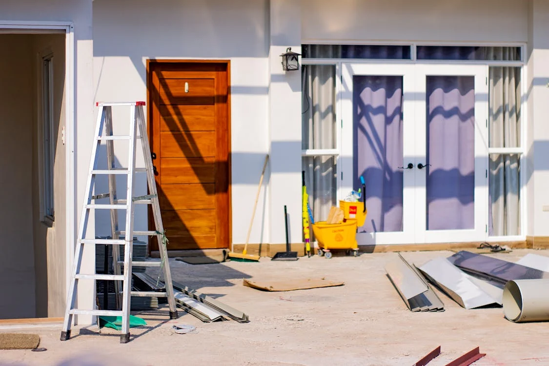 The image shows the exterior of a house under construction. There is a ladder leaning against the wall, a yellow cleaning cart with supplies, and various construction materials scattered on the ground. The house has a white exterior with a wooden door and a large window with a curtain. The scene is bathed in bright sunlight, casting long shadows from the objects in the foreground. The image suggests that the house is nearing completion, with the finishing touches being added. The overall impression is one of progress and anticipation.