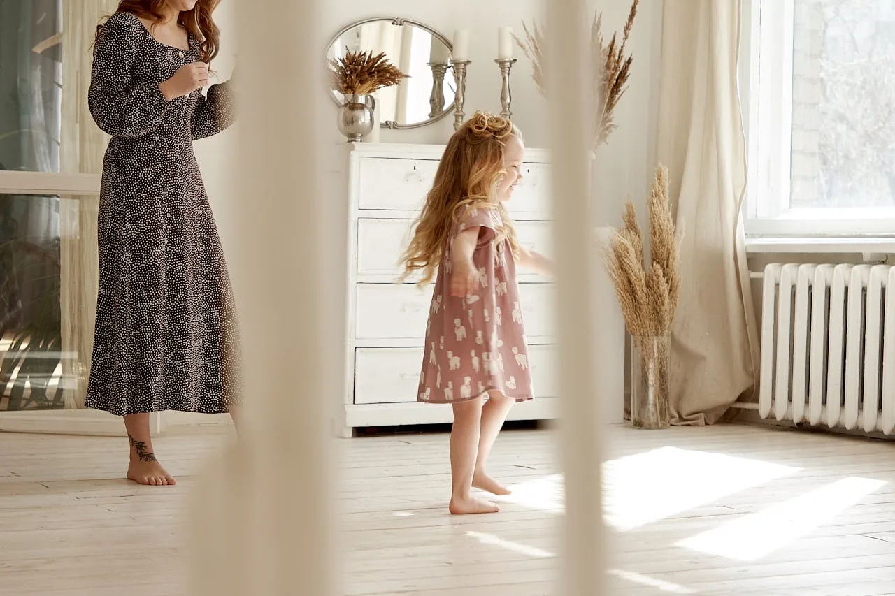 The image shows a young girl in a pink dress with a white pattern, spinning and laughing in a bright, airy room. She is barefoot and her long blonde hair is flowing behind her. A woman in a black and white polka dot dress is standing in the background, seemingly watching the girl with a smile. The room is decorated in a minimalist style, with white walls, wooden floors, and a few pieces of furniture. The light is soft and warm, creating a cheerful and playful atmosphere. The image captures a moment of pure joy and innocence, as the girl spins and laughs with abandon.